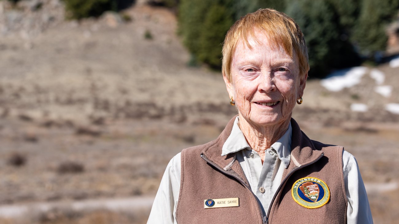 A park volunteer wearing a brown uniform vest over a khaki shirt smiles at us.